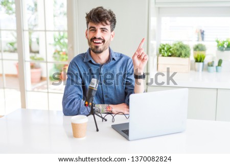 Young man recording podcast using microphone and laptop with a big smile on face, pointing with hand and finger to the side looking at the camera.