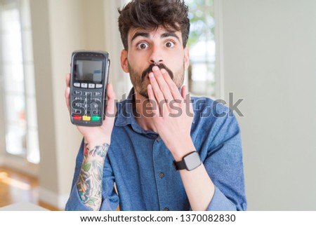 Young man holding dataphone point of sale as payment cover mouth with hand shocked with shame for mistake, expression of fear, scared in silence, secret concept