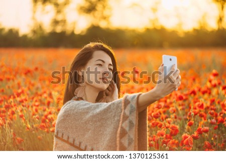 Smiling beautiful young woman boho stylish taking selfie portrait with smartphone in red poppies flowers meadow at sunset.