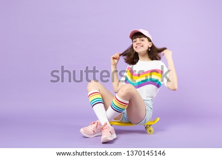 Beautiful teen girl in vivid clothes sitting on yellow skateboard, looking camera isolated on violet pastel wall background in studio. People sincere emotions lifestyle concept. Mock up copy space