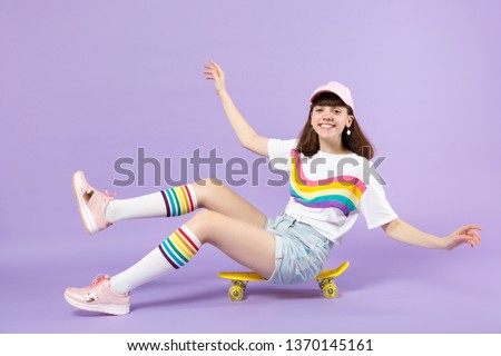 Funny cute teen girl in vivid clothes sitting on yellow skateboard, spreading hands isolated on violet pastel wall background in studio. People sincere emotions lifestyle concept. Mock up copy space