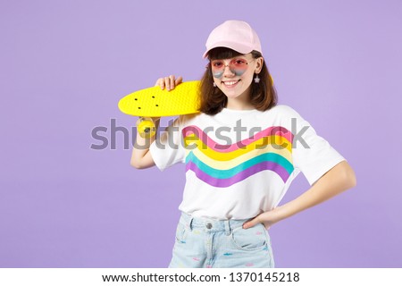 Portrait of beautiful teen girl in vivid clothes, heart eyeglasses holding yellow skateboard isolated on violet pastel wall background. People sincere emotions, lifestyle concept. Mock up copy space