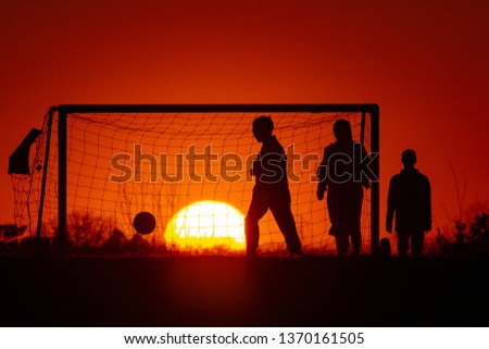 The silhouette picture of a few Children plays soccer at with the goal and a big sunset on the background.