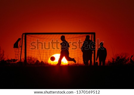 The silhouette picture of a few Children plays soccer at with the goal and a big sunset on the background.
