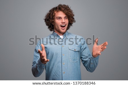 Handsome male in blue shirt gesturing with hands and looking away with astonished face expression while standing on gray background
