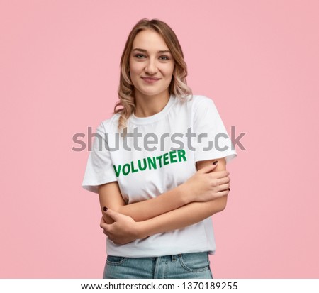 Positive young woman in white T-shirt with volunteer writing embracing herself and looking at camera while standing on pink background