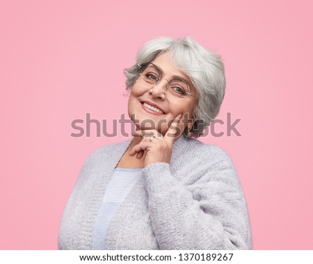 Young in heart elderly lady in stylish glasses cheerfully smiling and touching cheek while standing on pink background. Good looking old woman for nursing home