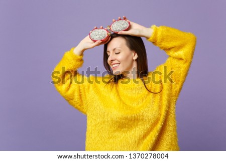 Relaxed young woman in fur sweater keeping eyes closed holding halfs of pitahaya, dragon fruit isolated on violet pastel background. People vivid lifestyle relax vacation concept. Mock up copy space