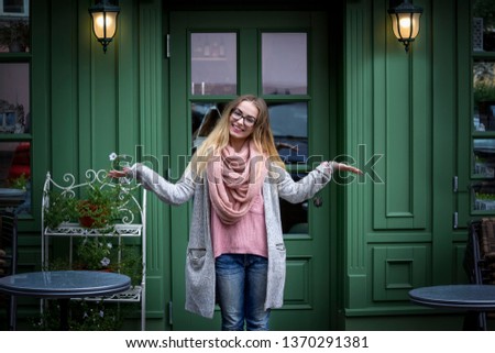 A sweet, young, long-haired blonde is standing by a trendy French-style cafe.