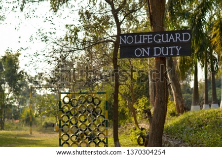 No life guard on duty signage wooden signboard in a hill resort with painted tires in green & yellow color arranged vertically to make an obstacle course to climb in a park in the background