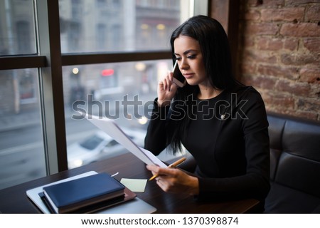 Young caucasian woman prosperous business worker holding paper document with monthly revenue report and talking with partner via cell telephone while sitting in restaurant. Manager reading resume 