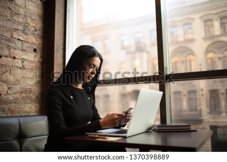 Woman prosperous lawyer online banking via mobile phone during work on laptop computer, sitting in modern restaurant interior. Female proud economist sending sms via cell telephone, relaxing in cafe 