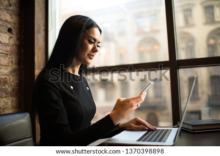 Smiling successful woman marketer received notifications on mobile phone during work on laptop computer, sitting in office space. Female government worker checking e-mail on cellphone during webinar