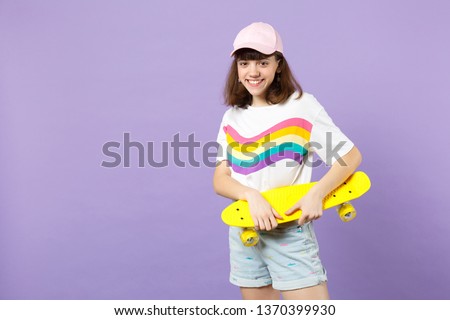 Portrait of pretty teen girl in vivid clothes looking camera, holding yellow skateboard isolated on violet pastel background in studio. People sincere emotions, lifestyle concept. Mock up copy space