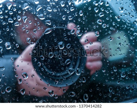 An elderly male photographer with beard and glasses is reflected in the mirror of car with close-up camera. On  sight of car large drops of rain. photographer is keen on filming in nature.