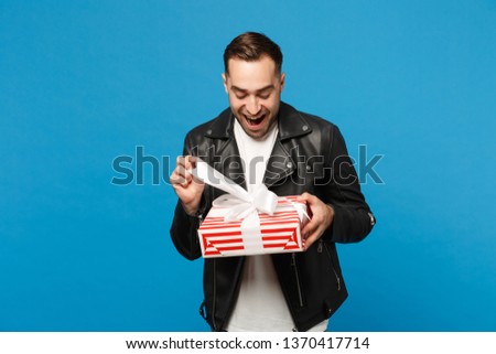 Handsome stylish young unshaven man in black leather jacket white t-shirt hold gift box isolated on blue wall background studio portrait. People sincere emotions lifestyle concept. Mock up copy space