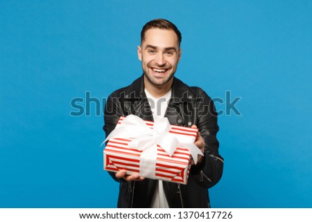 Handsome stylish young bearded man in black leather jacket white t-shirt hold gift box isolated on blue wall background studio portrait. People sincere emotions lifestyle concept. Mock up copy space