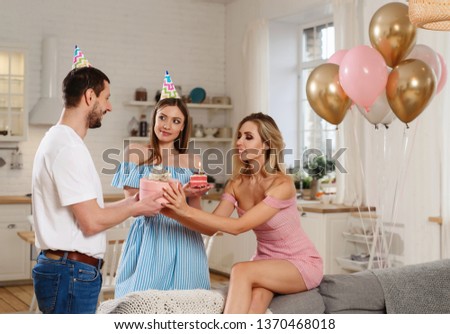 Beautiful happy Caucasian blonde woman blowing candles on birthday cake. Friends taking a gift to girl. Birthday party with friends concept