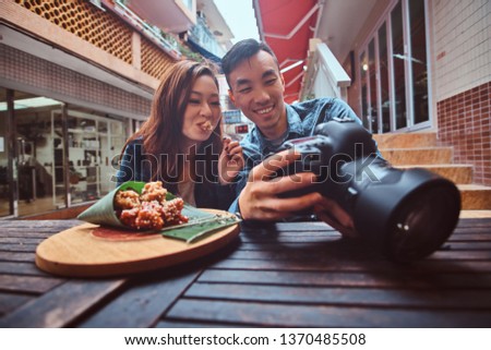 Happy young chinese couple sitting at cafe outside enjoying traditional asian food and watching pictures in photo camera. Widescreen shooting.