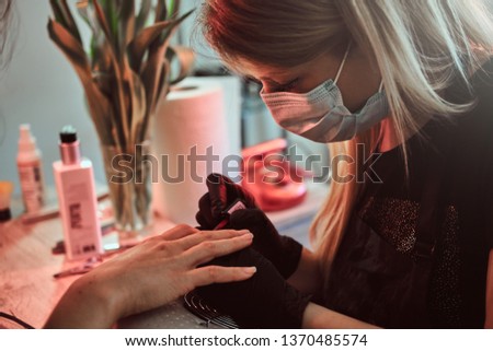 Young manicure master is working on client's nails. She is focused on client nails. Master is wearing a protection mask.