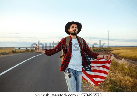 4th of July. Fourth of July. American with the national flag. American Flag. Independence Day. Patriotic holiday. The man is wearing a hat, a backpack, a shirt and jeans. Beautiful sunset light.  