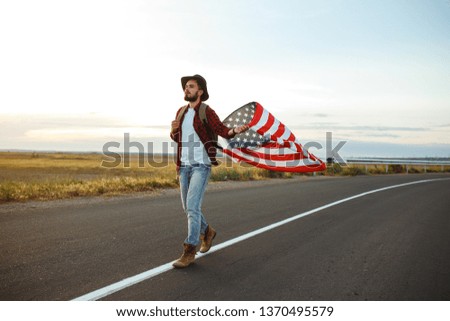 4th of July. Fourth of July. American with the national flag. American Flag. Independence Day. Patriotic holiday. The man is wearing a hat, a backpack, a shirt and jeans. Beautiful sunset light.  