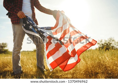 4th of July. Fourth of July. American with the national flag. American Flag. Independence Day. Patriotic holiday. The man is wearing a hat, a backpack, a shirt and jeans. Beautiful sunset light. 