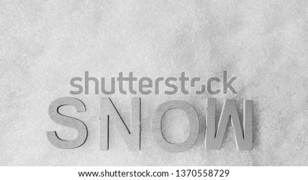 The word SNOW spelled out in capitalized wooden letters, isolated on a white background of fresh, real snow. Black and white, offset low, horizontal, closeup.