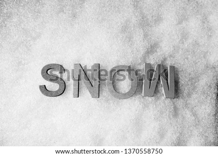 The word SNOW spelled out in capitalized wooden letters, isolated on a white background of fresh, real snow. Black and white, middle, horizontal, closeup.