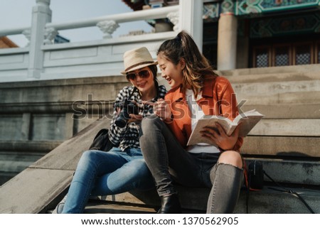 two happy young asian girls travelers with guided book sitting in stairs in chinese confucius temple background searching tourist destination. women point camera look check picture having fun chat