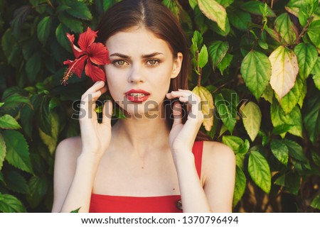 woman in a bright dress with a flower in her hair looks into the camera and stands near the bushes                          