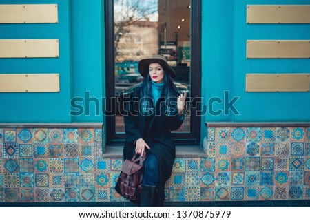 Beautiful young woman with blue hair in nice black coat, jeans and hat. High heels shoes. Spring fashion photo on urban background.