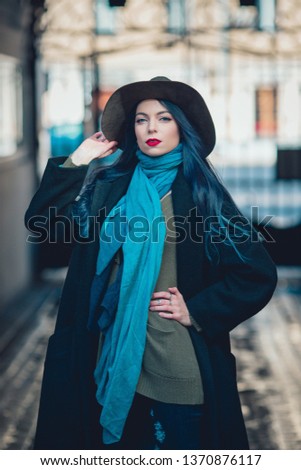 Beautiful young woman with blue hair in nice black coat, jeans and hat. High heels shoes. Spring fashion photo on urban background.