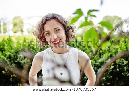 Portrait of young woman with red lipstick in white dress outdoor. Smiling woman. Curly brown hair girl in the summer park