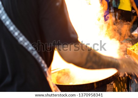 Out of focus of Fire burn during cooking on iron pan. Chef Cooking With Fire In Frying pan.Night picture of a street food chef cooking meat and fish in a pan with fire and flames under it.