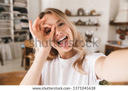 Image closeup of amusing blond woman 20s wearing white t-shirt smiling while looking at camera and taking selfie photo in living room