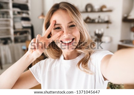 Image closeup of pleased blond woman 20s wearing white t-shirt smiling while looking at camera and taking selfie photo in living room