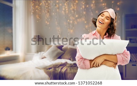 people and bedtime concept - happy young woman in pajama and eye sleeping mask hugging pillow over home bedroom background