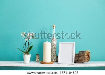 Modern room decoration with Picture frame mockup. White shelf against pastel turquoise wall with Candle and rocks in bottle. Potted orchid plant