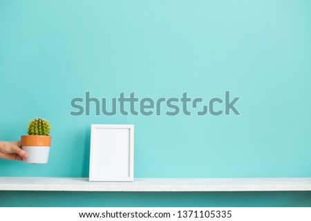Modern room decoration with Picture frame mockup. White shelf against pastel turquoise wall with hand putting down potted cactus plant.