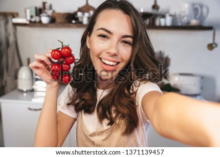 Attractive brunette woman wearing apron taking a selfie while sitting at the kitchen at home, holding bunch of tomatoes