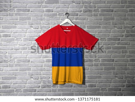 Armenia flag on shirt and hanging on the wall with brick pattern wallpaper, red blue and orange.