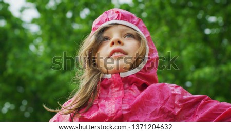 Authentic shot of cute happy little girl in raincoat is having fun and enjoying the rain in a hot summer day in a green park.