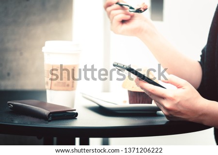 Closeup image of a woman holding , using and looking at smart phone while eating a cake in cafe shop