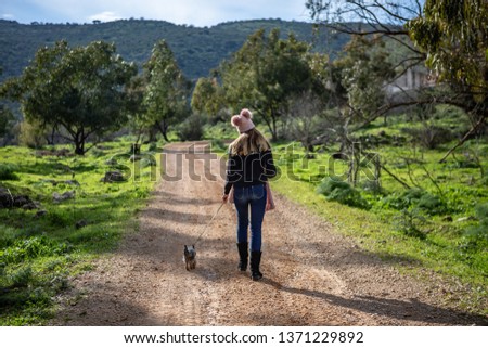 A girl in a pink hat with pom-poms and a scarf walks with a Yorkshire terrier on a country road to a ridge