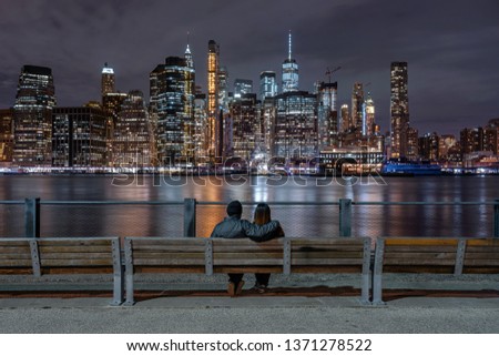 Scene of back side couple sitting and looking New york Cityscape beside the east river at the night time, USA downtown skyline, Architecture and building with lover concept