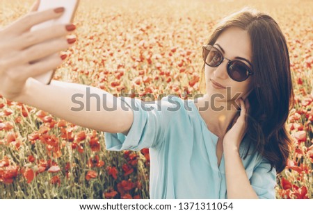 Pretty stylish young woman taking selfie with smartphone in poppies flowers meadow in spring outdoor.