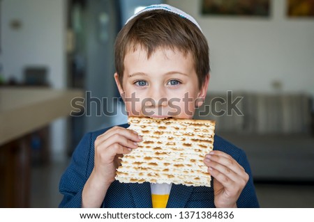 Cute Caucasian Jewish boy holding in his hands and taking a bite from a traditional Jewish matzo unleavened bread. Jewish Passover Pesach concept image. Royalty-Free Stock Photo #1371384836