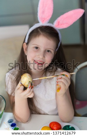 Beautiful little brunette girl, has happy fun smiling face, pretty eyes, white t-shirt, hare ears, paint Easter eggs. Child portrait and kids hobby concept. Holiday accessories. 