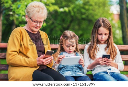 Family alienation. Grandmother sitting in the park and using smartphone while her granddaughters have fun with mobile phone and tablet. Family, lifestyle, technology concept
